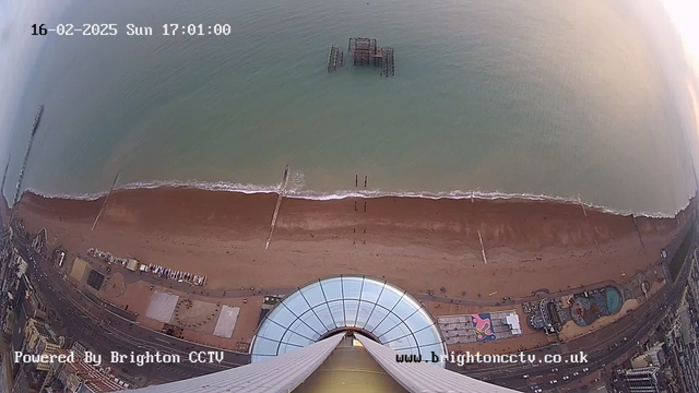 An aerial view of Brighton beach, showing a wide expanse of sandy shore meeting the bright blue sea. There are people walking along the beach, with a pier visible extending into the water from the left side of the image. The lower part features buildings and attractions, including a circular structure near the front. The sky above is clear with a hint of sunset colors. A timestamp indicating the date and time is located at the top of the image.