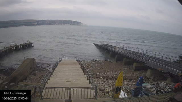 A view of the sea under a cloudy sky. In the foreground, there are stone steps leading down towards the water, flanked by railings. To the left, there is a wooden jetty extending into the water. A rocky shoreline is visible, with a few boats and kayaks in bright colors (yellow, blue, and red) resting on the ground. In the distance, a cliff can be seen, and the water appears slightly choppy with small waves.