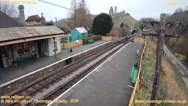 A railway station platform with empty tracks extends into the distance. On the left, there is a stone building with a peaked roof and tall chimney, featuring a green bench and a sign on the wall. A blue shed is partially visible behind a wooden fence that has a small "Way Out" sign. Several green benches line the platform, while a single person stands near the edge of the tracks. In the background, a castle ruins are perched on a hill, surrounded by trees and a cloudy sky.