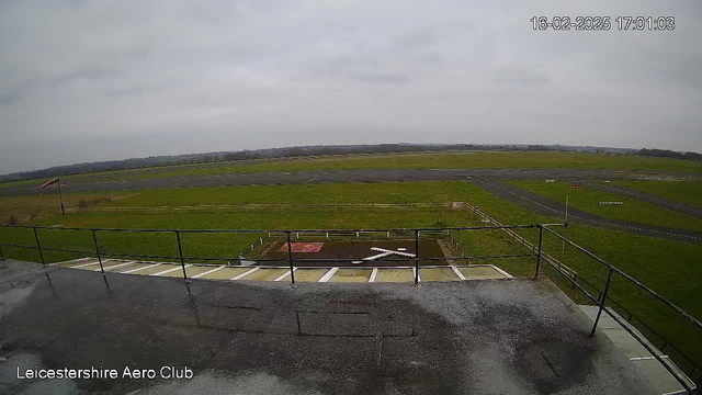 A wide view of an open field with a runway, captured from a balcony of a building. The ground is mostly green grass, with some dirt patches. A white cross is visible on the ground in front of the balcony. In the background, there are distant fields and a cloudy sky, indicating overcast weather. A windsock is positioned on the left side of the image, and the area is fenced. The scene feels spacious and open, typical of an airfield. The time and date are displayed in the upper right corner.