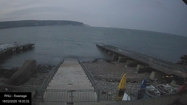 A view of a rocky shore with a calm sea. In the foreground, there is a wide set of stairs leading down to the water, bordered by a metal railing. To the right, a small dock extends out into the sea. Two kayaks, one yellow and one blue, are stored alongside the steps. In the background, a cliff is visible along the edge of the coastline, and the sky appears overcast, indicating early evening. The scene is tranquil with gentle waves lapping at the shore.