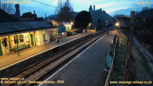 A railway station platform is depicted in the image, with a partially illuminated station building on the left. The platform features green benches and a notice board. In the background, there are tracks leading into the distance. A medieval castle can be seen on a hilltop in the far background, surrounded by trees. The sky is at dusk, with a bluish tint. There is also a wooden utility pole on the right side of the image, with wires attached.