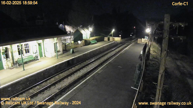 A nighttime view of a railway station platform. On the left, there is a building with a light illuminating a bench and some signage. The platform is mostly empty with a green bench, and there are two railway tracks running parallel along the bottom of the image. In the background, there are faint lights from nearby trees and the outlines of fences. A sign reading "MIND OUT" is positioned on the ground near the tracks. The setting is quiet and sparsely lit, indicative of a serene night atmosphere.