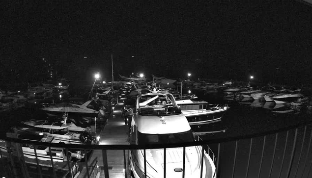 A nighttime scene at a marina filled with boats. The image is predominantly dark, with only a few lights illuminating parts of the dock and surrounding boats. Several vessels are moored closely together, and a wooden walkway leads deeper into the marina, surrounded by water reflecting faint light. The boats vary in size and shape, with some appearing larger and others smaller, but all are partially obscured by shadows.