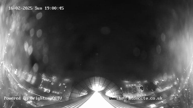 A black and white webcam image showing a cityscape at night. The lower portion features bright lights from vehicles or buildings, while the upper section is mostly dark, with some light reflecting off a surface. The time stamp at the top reads "16-02-2025 Sun 19:00:45" and there is a watermark for Brighton CCTV at the bottom. The image appears slightly hazy with some light reflections.