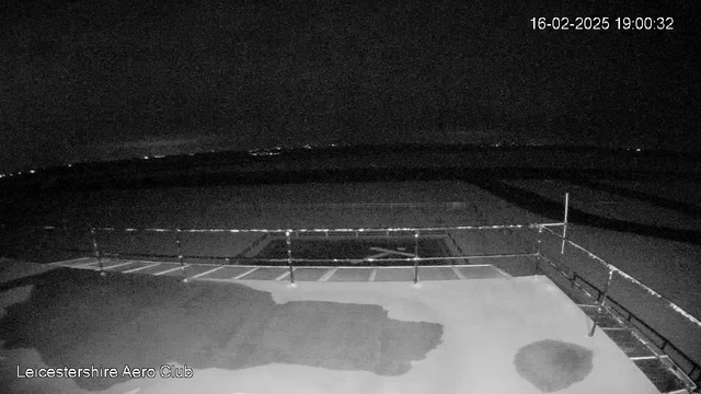 A dark scene captured by a webcam, featuring the outline of a runway at the Leicestershire Aero Club. The foreground shows a flat surface with some water patches, while a railing borders the area. In the background, dim lights hint at distant locations, and the sky is mostly dark with little visible detail. A timestamp indicates the image was taken on February 16, 2025, at 7:00 PM.