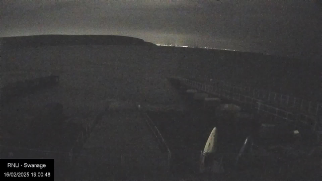 A dark image of a coastal scene. In the foreground, there are wooden docks and some boats. The water appears calm, reflecting a cloudy, dimly lit sky. In the background, a distant shoreline with faint lights can be seen, indicating a town or settlement. The overall atmosphere is serene and quiet. The timestamp at the bottom indicates the date and time.