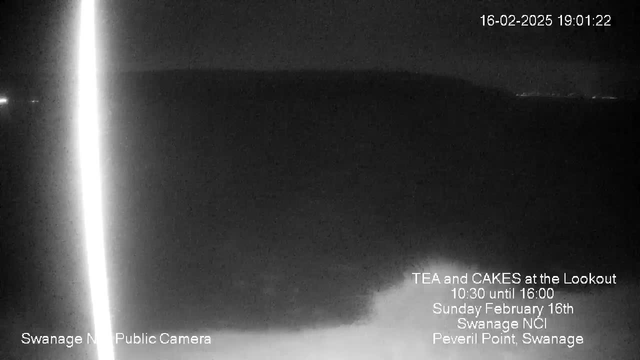 A dark, nighttime scene captured from a webcam. The horizon is barely visible against a black sky, with faint outlines suggesting land in the distance. On the left side, there is a bright vertical light reflecting off the water. In the lower part of the image, text messages provide information about a local event, indicating that tea and cakes will be served at the Lookout on Sunday, February 16th, from 10:30 to 16:00. The bottom left corner displays the camera's location as "Swanage NCI Public Camera."