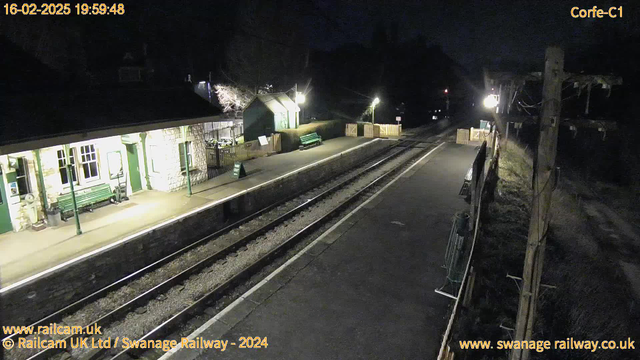 A dark scene at a train station at night. The platform is empty, with a few benches visible. There is a stone building with green trim and windows on the left side, which appears to be the station house. On the platform, there is a sign leaning against a post that reads "WAY OUT." There are two benches along the platform and a low wooden fence. In the background, there are dim lights from the station and a distant light from a nearby building. The railway tracks are visible, leading off into the darkness.