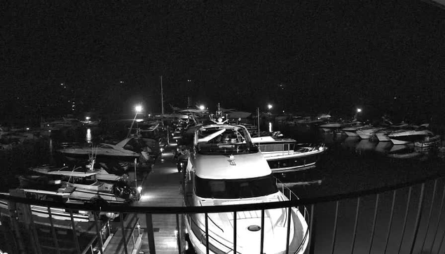 A nighttime scene at a marina, featuring multiple boats docked in the water. The image is predominantly dark, with only a few lights illuminating the area. A large boat is in the foreground, with a visible deck and railing. Surrounding boats are mostly obscured but are arranged closely together in the water. Some of the boats are equipped with navigation lights. The atmosphere is quiet and serene, typical of a nighttime harbor setting.