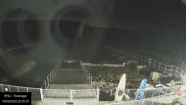 A dark, nighttime image of a waterfront area. In the foreground, a wooden dock extends into the water, bordered by white railings. On the right, two brightly colored surfboards lean against the railing. The ocean is visible, with gentle waves lapping against the shore, and dark rocky areas are seen beneath the water. The background is a dark sky with distant lights possibly indicating a city or urban area far away.