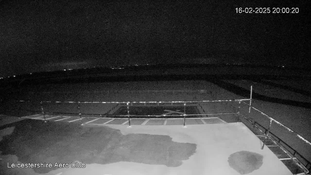 A dark scene captured from a webcam overlooking a flat surface with faint outlines of a runway or tarmac below. The ground appears wet in patches, possibly from rain. In the distance, faint lights can be seen, likely from far-off buildings or vehicles. There is a railing in the foreground, and the image has a timestamp indicating the date and time. The setting suggests it is early evening or night.