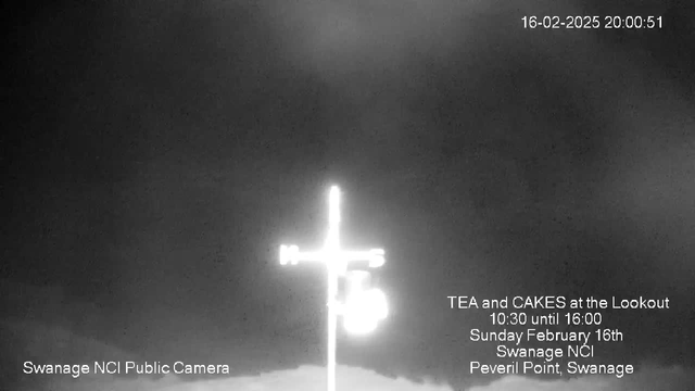 A black-and-white webcam image featuring a silhouette of a cross atop a pole at Peveril Point, Swanage. The background shows a cloudy sky, and text on the image indicates the date and time as February 16, 2025, at 20:00:51, along with an announcement for a tea and cakes event at the lookout from 10:30 to 16:00. The bottom of the image displays the name "Swanage NCI Public Camera."