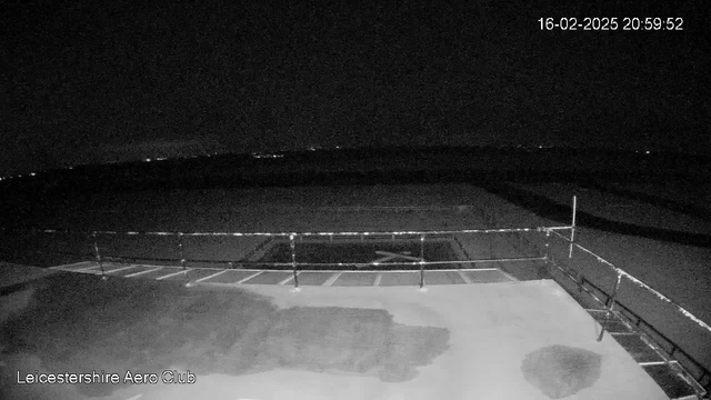 A dark, nighttime image captured from a webcam, showing a scene of an airfield. In the foreground, there's a flat, manmade surface with some markings and patches of light. A low fence with horizontal bars runs along the edge of the surface, and in the distance, faint lights suggest a landscape beyond the airfield. The overall atmosphere is calm and quiet, indicative of late evening with no visible activity. The timestamp on the image indicates it was taken on February 16, 2025, at 20:59:52.