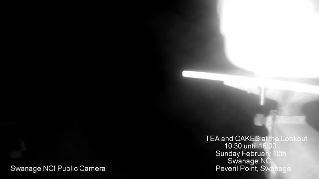 A black and white webcam image showing faint outlines of a light source, with bright highlights and dark shadows surrounding it. Text in the lower left corner reads "Swanage NCI Public Camera," and text on the right side provides event details: "TEA and CAKES at the Lookout, 10:30 until 16:00, Sunday February 16th, Swanage NCI, Peveril Point, Swanage." The overall image appears to be low light with unclear visibility.