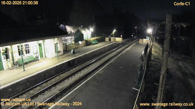 A dimly lit railway station platform at night, featuring a stone building with large windows and green accents. There are benches along the platform, and a green sign reading "OUT" is visible. The railway tracks run parallel to the platform, and a wooden picket fence is seen in the background. Nearby, another bench is next to a green shed or building, with limited lighting illuminating the area. The scene conveys a quiet and isolated atmosphere.