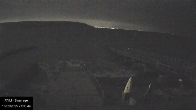 A dark landscape is captured, with a faint horizon in the background indicating distant lights. In the foreground, there are vague shapes of objects, possibly boats or equipment, along with a wooden structure or pier extending towards the right. The scene appears to be nighttime, with low visibility and a calm ambiance.