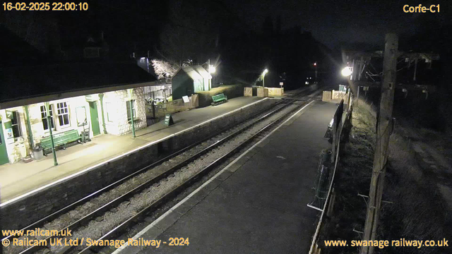 A dark scene showing a train station at night. The platform is mostly empty with a few benches on the left and a sign that reads "WAY OUT." The station building features stone walls and green trim, with several windows lit up. On the right side, there are two sets of railway tracks running parallel, leading off into the darkness. Lights from the station and a nearby signal illuminate parts of the scene, creating a stark contrast with the surrounding night.