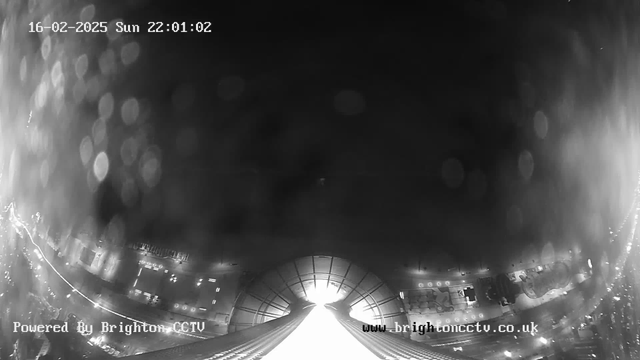 A black and white surveillance image showing a view from a high vantage point. The scene appears dimly lit with reflections and a bright light source in the background, possibly indicating a street or building entrance. There are blurred shapes and lights indicating structures below, with some light trails suggesting movement. The date and time are displayed in the upper left corner, and a watermark for Brighton CCTV is present in the lower left corner.