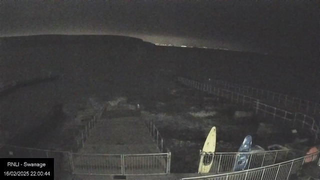 A dimly lit scene of a shoreline at night, with a faint outline of a hill in the background. In the foreground, there are two kayaks, one yellow and one blue, positioned near a fenced area. The water appears dark and still, with some small waves visible. The sky is cloudy, obscuring most stars, with a faint glow on the horizon. The date and time display indicates it is February 16, 2025, at 10:00 PM.