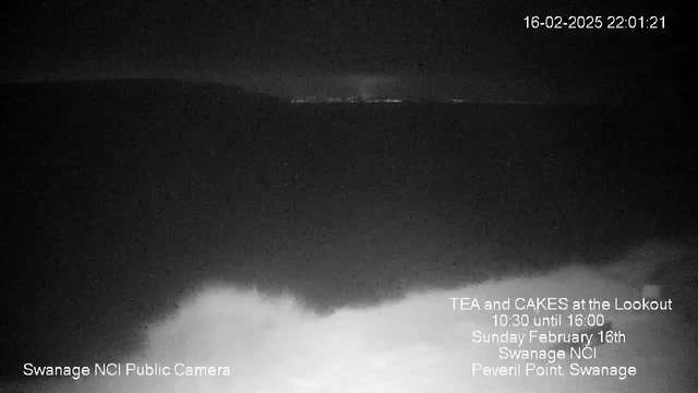 A dark image showing a cloudy night sky with low visibility. In the distance, faint lights are visible along the horizon, indicating a distant land or settlement. The bottom of the image features a rough, white area, hinting at the presence of waves or fog. Text in the lower right corner announces an event, "Tea and Cakes at the Lookout," with details of the date and location.