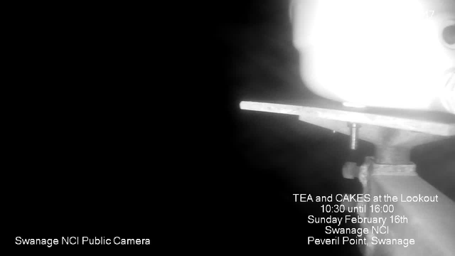 A dark image featuring a blurred, illuminated object on the right side, likely a camera or light source. The background is predominantly black. Text on the bottom left reads "Swanage NCI Public Camera," and on the right side, there is additional text advertising a "Tea and Cakes" event at the Lookout, including the date and time: "10:30 until 16:00, Sunday February 16th, Swanage NCI, Peveril Point, Swanage."