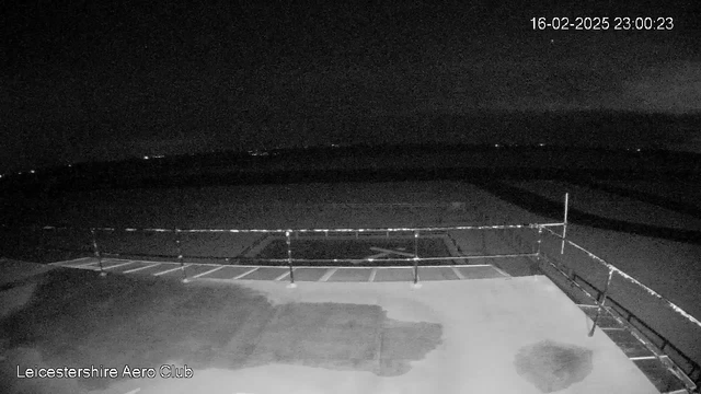 A dark scene from a webcam showing the Leicester Aero Club at night. In the foreground, there is a flat surface with a faint outline of what could be parking spaces. A railing is visible along the edge, and there are indistinct lights in the distance indicating possible structures or vehicles. The sky is mostly dark with subtle variations in shade, suggesting cloud cover. The timestamp in the corner indicates February 16, 2025, at 23:00:23.