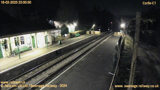 A dimly lit railway station at night. The foreground shows two parallel railway tracks running through the center. On the left, there is a platform with a low, stone wall and wooden benches painted green. A small building with large windows is visible, partially illuminated by lights. To the right of the building, there is a green sign that says "OUT" mounted on a wooden fence. Several of the benches are empty, and the surrounding area is dark with faint overhead lighting. Trees are visible in the background, adding to the nighttime atmosphere.