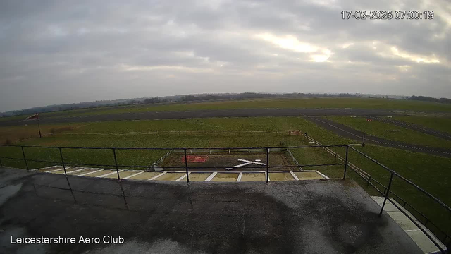 A view from the rooftop of a building at the Leicestershire Aero Club. The foreground shows a flat, dark surface with a railing along the edge. Below the railing, there is a grassy area and an airstrip with a small grassy patch. A white cross is visible on the ground, likely marking a landing spot. In the background, the landscape features a grey, overcast sky with clouds, and a distant horizon with a few trees. The time stamp in the upper right corner indicates the date and time of the image.