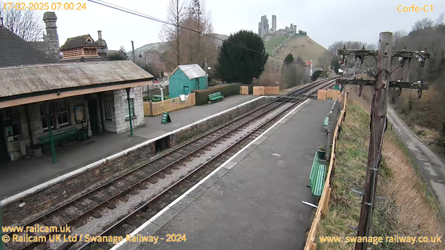 A scenic view of a train station with a stone building on the left and green benches along the platform. In the background, there are rolling hills, and on the hill, remnants of a castle can be seen. The platform has a few wooden fences, and a green sign indicating "Way Out" is positioned near the station's edge. The tracks run parallel to the platform, leading off into the distance. The scene is set in a cloudy atmosphere, and there are trees lining the background.