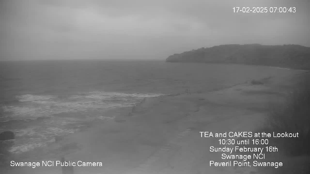 A grayscale image of a cloudy beach scene. The sea is visible with gentle waves lapping at the shore. A rocky coastline extends in the distance to the right, partly obscured by mist. The sky is overcast, and the overall atmosphere is tranquil yet somber. In the lower left corner, there is text that indicates the camera location: "Swanage NCI Public Camera." In the lower right corner, additional information is provided about an event, including times and dates, but it is also obscured by the cloudiness of the image.