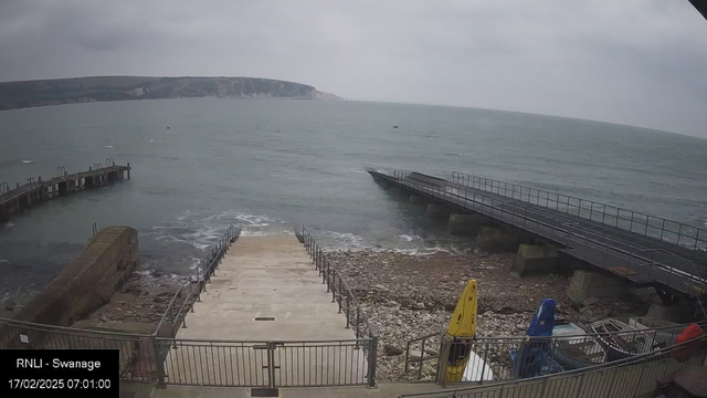 A coastal scene showing a sweeping view of the sea under a cloudy sky. To the left, a pier extends into the water, with gentle waves lapping against it. In the foreground, there is a set of concrete steps leading down to the water's edge, flanked by a metal railing. Scattered stones are visible along the shore. On the right side, there are several colorful kayaks positioned on the ground near a wooden structure. The overall atmosphere is tranquil and slightly overcast.