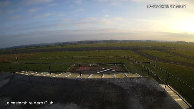 A view from a webcam at the Leicestershire Aero Club, showing a wide grassy landscape under a partly cloudy sky. In the foreground, there is a railing and a rooftop surface. A white "X" symbol is marked on the ground, surrounded by a fence. The background features an airstrip and open fields, with distant hills visible under soft lighting from early morning.