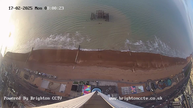 A wide, aerial view of a beach with brown sand and gentle waves lapping at the shore. An abandoned pier is visible in the water, along with several small wooden posts extending into the sea. To the left, a portion of a boardwalk and colorful amusement park with a circular pool can be seen. The sky is partly cloudy, and the image is timestamped at 08:00:23 on February 17, 2025. The bottom of the image shows the top of a tall structure pointing downwards, along with the text "Powered By Brighton CCTV" and a website link.