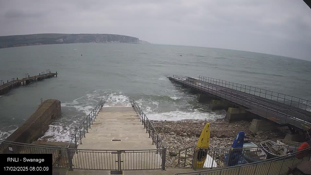 A cloudy sky over a calm sea with gentle waves. In the foreground, there are stone steps leading down to the water, bordered by a metal railing. To the left, a small concrete pier extends into the sea, and to the right, a larger, flat dock is seen, both with railings. On the shore, some kayaks in bright yellow and blue are positioned near the water's edge, along with some scattered rocks. The scene conveys a serene coastal setting.