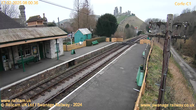 A view of a train station with a stone building featuring a sloped roof and large windows. There is a green bench on the platform, and a small teal shed is visible in the background. A wooden fenced area has two green benches and a sign that says "RAY OUT." Two railway tracks extend into the distance, and in the background, there are hills with ruins, suggesting a castle. The sky is overcast and the scene appears quiet and rural.