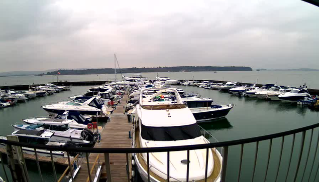 A panoramic view of a marina filled with various boats and yachts. The water is calm and reflects the overcast sky. There are several wooden docks visible, with groups of boats moored alongside them. In the background, a shoreline is faintly visible with trees and hills. The scene has a quiet and serene atmosphere, typical of a cloudy day at a harbor.