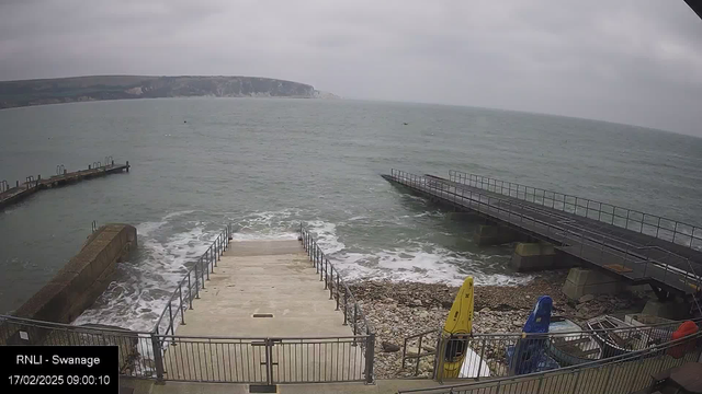 A view of a coastal area with a gray sky and gentle waves. In the foreground, there are stone steps leading down to the water. To the right, a wooden jetty extends into the sea, and to the left, a curved stone wall protects a section of the beach. There are several brightly colored kayaks, one yellow and one blue, positioned near the edge. The sea appears calm, with small ripples and a distant horizon where land meets the water.