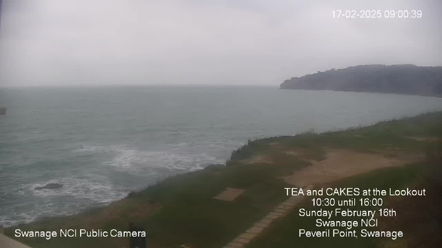 A cloudy sky is visible above a calm sea, with gentle waves lapping at the shore. The grassy area to the right shows a path leading towards the water. In the distance, a cliff rises from the coastline. Text overlays at the bottom mention a public camera in Swanage, details about a tea and cakes event, and the date and time.