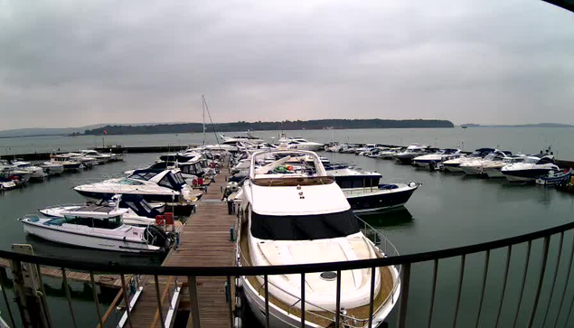 A view of a marina on a cloudy day, featuring multiple boats docked in the water. The foreground shows a wooden pier with several boats moored alongside it, including a large white yacht. In the background, more boats are visible, and the water appears calm. A distant shoreline with trees can be seen against the overcast sky.