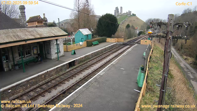 A view of Corfe Castle Railway Station on a cloudy day. The platform features stone buildings with a sloped roof, green benches, and a sign that reads "BAY OUT." To the left, there is a small green building, and in the background, Corfe Castle is visible atop a hill. The railway tracks run parallel along the platform, with a wooden fence and trees lining the area. A telegraph pole stands on the right, and a train is seen approaching in the distance.