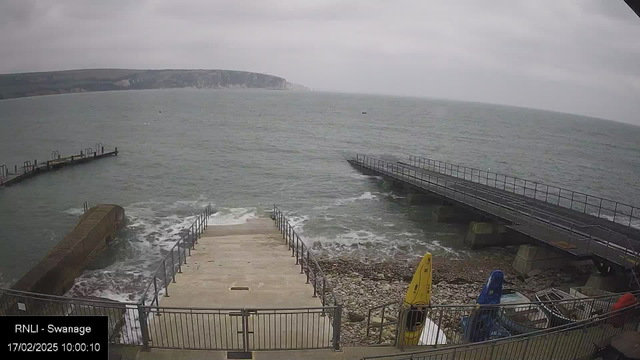 A cloudy scene featuring a coastline. The ocean is visible, with gentle waves lapping at the shore. A set of concrete steps leads down to the water, flanked by a railing. On the right, there is a rocky area with a mix of stones and pebbles. Two kayaks, one yellow and one blue, are positioned on the ground. In the background, a pier extends into the water with a railing. The sky is overcast, contributing to a muted color palette.