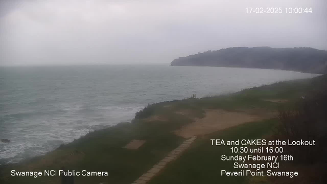 A cloudy seascape with a grayish sky and choppy greenish-blue water. In the foreground, there is a grassy area with a path made of stones leading towards the water. On the right, a rocky coastline rises, partially obscured by mist. Text in the lower corner indicates it is a public camera from Swanage NCI, and additional text highlights a tea and cake event at a lookout point, with a date and time displayed.