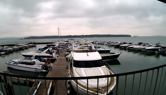 A view of a marina with numerous boats and yachts moored along a wooden dock. The water is calm and reflects the overcast sky, which is gray and cloudy. In the background, there are hills or land visible across the water. The scene conveys a serene maritime environment.