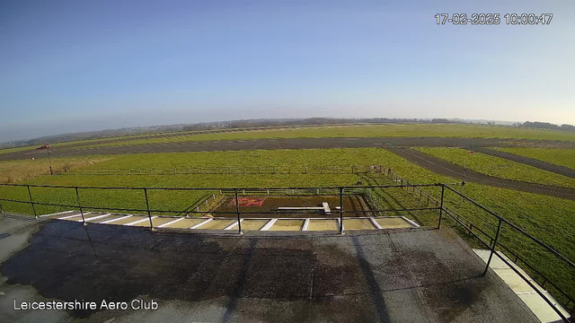 A view from a webcam situated on a rooftop, overlooking a large, open area of grass and an airstrip. The sky is clear and blue. In the distance, there are low hills and a few scattered trees. The foreground features a metal railing and a concrete surface. There are several markings on the ground, indicating sections of the airstrip, and a small structure near the bottom center of the image.