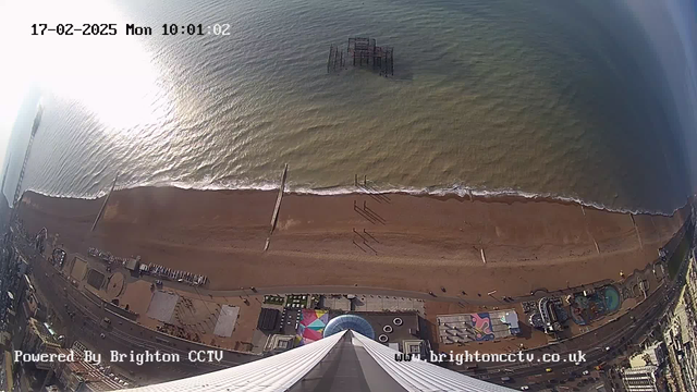 A view of a beach and ocean from a high vantage point. The sandy beach stretches horizontally across the image, with gentle waves lapping at the shore. In the foreground, there is a tall structure pointing downward, likely a tower. Some remnants of a pier can be seen in the water. Various people walk along the beach and the boardwalk, with a few colorful structures and amusement rides visible to the right. The sky is bright with sunlight reflecting off the water, creating a shimmering effect. The date and time are displayed at the top of the image.