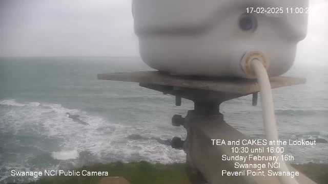 A coastal scene is shown with turbulent water waves crashing against the rocky shore. The focus is on a white cylindrical object, possibly part of a webcam setup, situated on a metal pole, overlooking the sea. The sky appears overcast and gray, indicating a cloudy day. Text in the lower portion provides information about a tea and cakes event at the Lookout, with details about time and date. The location is identified as Peveril Point, Swanage, and a logo indicates it is a public camera operated by Swanage NCI.