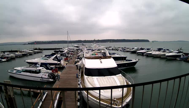 A view of a marina on a cloudy day. Several boats are moored in the water, including a large white yacht at the forefront with a tan top. Wooden docks extend into the water, and more boats are visible in the background. The sky appears overcast, and the shoreline can be seen in the distance.