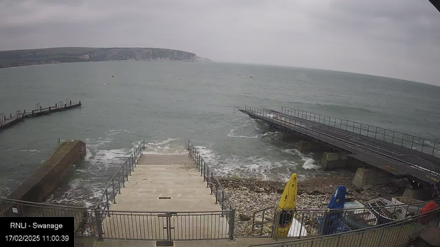 A cloudy coastal scene showing the sea with gentle waves. On the left, a wooden pier extends into the water, while on the right, a short set of steps leads down to the shore. There are scattered rocks and pebbles around the steps. Brightly colored kayaks in yellow, blue, and orange are positioned near the steps. The background features a rocky coastline with cliffs visible far in the distance.