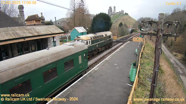 A train is departing from Corfe Castle station. The train is green, with a rounded nose and multiple windows along its side. In the background, there is a hill with ruins of a castle atop it, and several trees are visible. The station features a stone building with a sloped roof and a few benches along the platform. Overhead electrical wires and a wooden pole are also present, with nearby pathways and grassy areas. The sky appears cloudy.